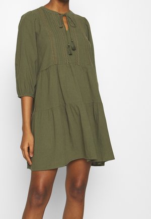 Vrouw draagt een loszittende olijfgroene jurk met driekwartmouwen, kantdetails en touwtjes met kwastjes bij de halslijn.