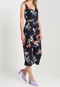 Marinblå blommig jumpsuit med v-ringning, ärmlös design och insydd midja. Har färgglada blommönster och når till mitt på vaden.