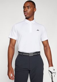 Hvit polo skjorte med krage, med en svart logo på venstre bryst. Svart belte med teksturerte detaljer og mørke bukser. Hvit golfhanske holdt i hånden.