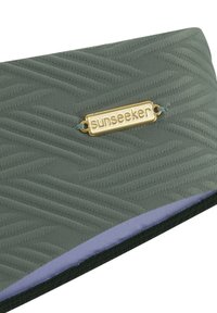 Zaļš rakstains audums ar teksturētu viļņu dizainu, kuram ir zelta "sunseeker" logo plāksne un violeta iekšējā mala.