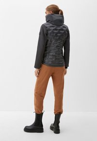 Frau, die von hinten blickt, trägt eine schwarze Daunenjacke, braune Jogginghosen und schwarze klobige Stiefel vor einem schlichten weißen Hintergrund.