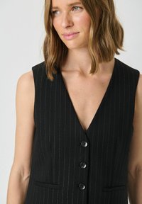 Black pinstripe vest with a deep V-neck, sleeveless design, three buttons, and two front pockets; textured fabric with subtle sheen.