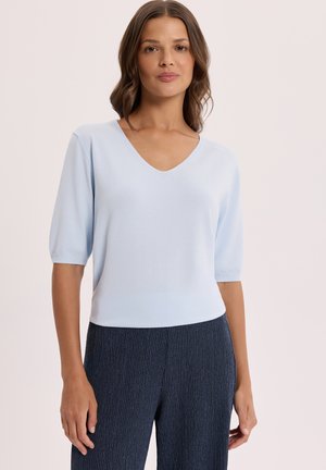 Kobieta ubrana w jasnoniebieską bluzkę z krótkim rękawem i dekoltem w serek oraz teksturowane ciemnoniebieskie spodnie, stojąca na tle jednolitego jasnego tła.