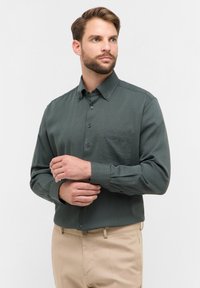 Donkergroene overhemd met knopen en een gestructureerde afwerking, lange mouwen, een zak op de linkerborst en bijpassende knopen. Gepaarde met beige broek.