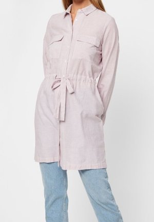 Kobieta w jasnoróżowej sukience koszulowej z długim rękawem, posiadającej przednie kieszenie i pasek w talii, w połączeniu z jasnoniebieskimi dżinsami.