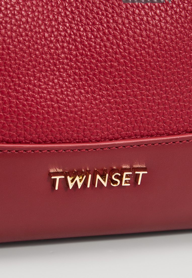 Superficie in pelle rossa testurizzata con rifiniture in pelle liscia e lettere metalliche dorate che formano la parola "TWINSET" su una borsa rossa.