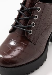 Chaussure à lacets en cuir verni brun, présentant un motif crocodile texturé, des lacets noirs, des œillets en argent et une semelle plateforme noire épaisse.