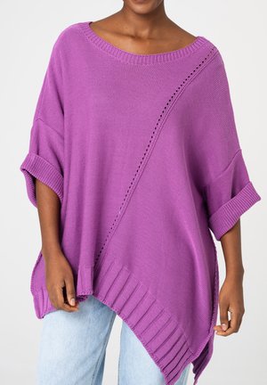 Kobieta ubrana w luźny fioletowy dzianinowy ponczo z podwiniętymi rękawami i prążkowanym dołem, w połączeniu z jasnoniebieskimi dżinsami, na jednolitym tle.