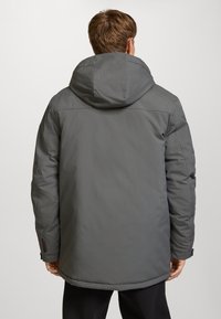 Chaqueta exterior gris con capucha, que presenta una textura suave, mangas largas y un dobladillo trasero ligeramente más largo. Costuras y herrajes detallados visibles.