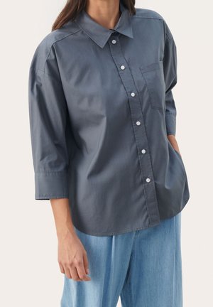 Frau trägt ein lockeres, dunkelgraues Hemd mit Knöpfen und dreiviertel Ärmeln sowie hellblaue Hose, Hand in der Tasche vor weißem Hintergrund.