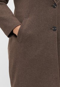 Casaco de lã castanho com um acabamento texturizado, apresentando uma única fileira de botões pretos e um bolso lateral, exibindo um design clássico.