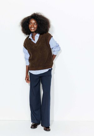 Gilet marron texturé sur une chemise à col bleu clair, associé à un pantalon large à rayures bleu marine et des chaussures foncées. Se tenant contre un fond blanc.