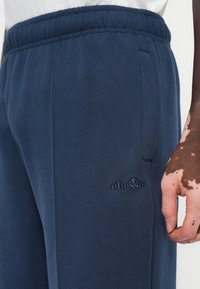 Temno modri trenirki iz mehkega blaga, z elastičnim pasom in stranskimi žepi. Logotip blagovne znamke je izšit na stegnu.