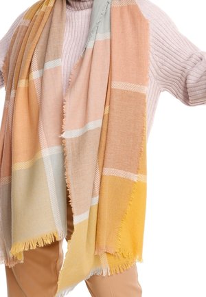 Personne portant un pull rose pastel et un pantalon beige, enveloppée d'une grande écharpe douce aux motifs à carreaux jaunes, pêche et crème.