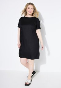 Schwarzes kurzärmliges Kleid aus weichem Stoff, mit lockerer Passform und rundem Ausschnitt. Tragen mit weißen Sandalen, die eine schwarze Sohle haben.