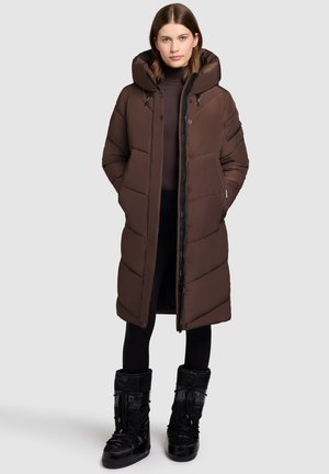 Vrouw in een lange bruine gewatteerde jas met capuchon, zwarte leggings en zwarte winterboots, staande tegen een effen lichte achtergrond.