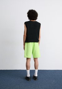 Haut en tricot sans manches noir et short vert fluo. Des chaussures noires et des chaussettes blanches complètent la tenue. Vue de dos sur un fond uni.