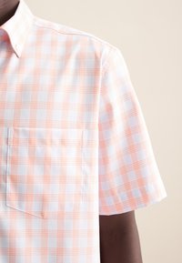 La camicia a maniche corte presenta un tessuto bianco con un motivo a scacchi arancione chiaro e blu, una tasca sul petto e un colletto button-down.