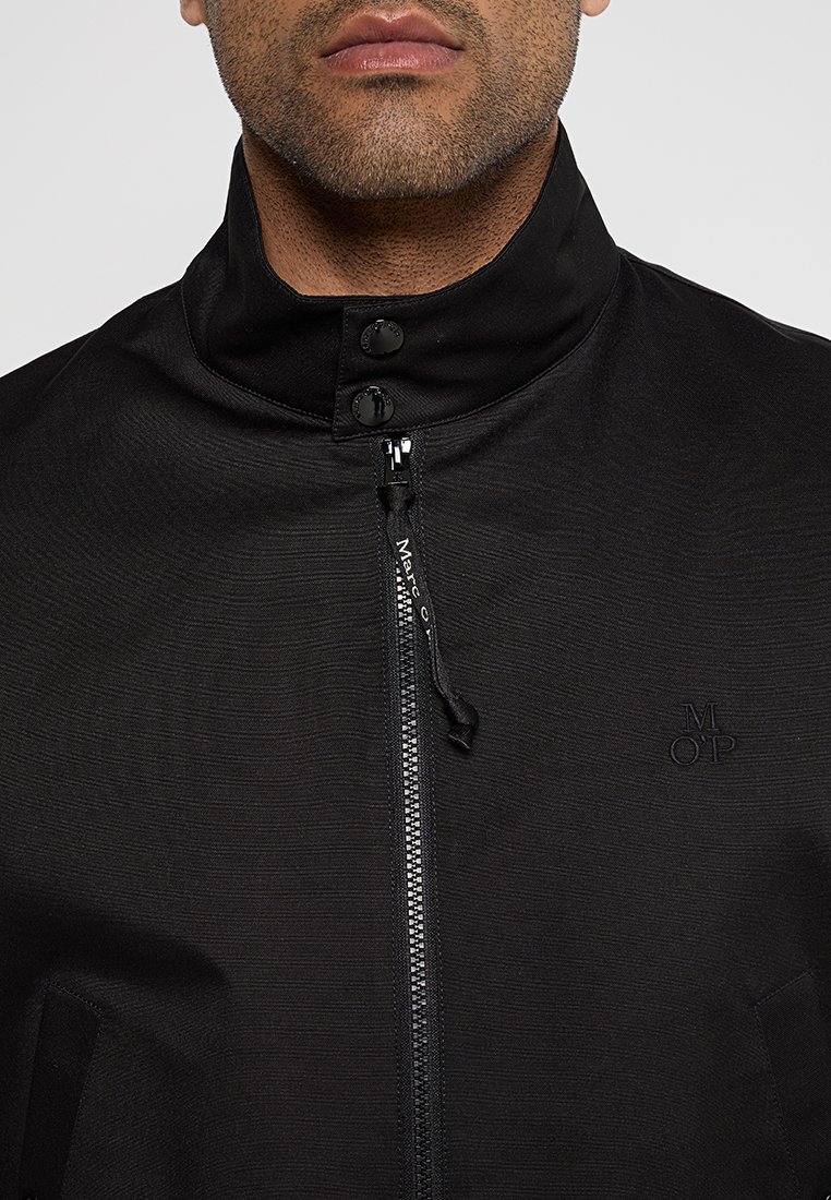 Schwarze Zip-Jacke aus strukturiertem Stoff mit hohem Kragen und dezentem Logodetail. Verfügt über einen Frontreißverschluss und Druckknöpfe.