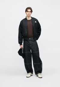 Schwarze Bomberjacke mit rundem Kragen, kombiniert mit lockeren dunklen Denimhosen mit weißen Nähten. Accessoires beinhalten eine schwarze Kappe.