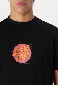 Black cotton t-shirt featuring a round pink graphic with "SANTA CRUZ" in orange lettering and a brick pattern background.