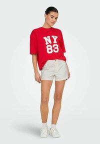Camiseta de algodón roja con gráfico "NY 83", combinada con pantalones cortos beige claros y zapatillas blancas. El modelo está de pie con las manos en las caderas frente a un fondo neutro.