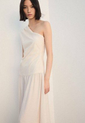 Femme aux cheveux courts et foncés portant une robe sans manches crème bustier, debout devant un fond clair uni.