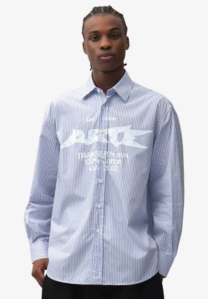 Giovane con i capelli intrecciati che indossa una camicia a righe blu e bianche con bottoni e testo bianco stampato sul davanti.