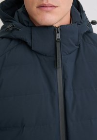 Marineblauwe pufferjas met een hoge kraag, ritssluiting aan de voorkant en verstelbare trekkoorden. Soepele stoftextuur met horizontale stikpatronen.