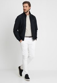Chaqueta bomber negra, camiseta de color gris claro, pantalones blancos y zapatillas negras. La chaqueta cuenta con un cierre de cremallera y bolsillos laterales.