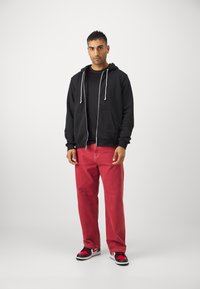 Sudadera negra con cremallera y cordones, camiseta negra, pantalones rojos y zapatillas negras/rojas. Tejido suave y corte holgado. Diseño simple.