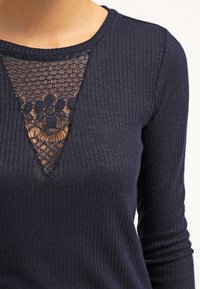 Haut à manches longues côtelé bleu marine avec un panneau en dentelle triangulaire au niveau de l'encolure, ajoutant texture et détail au design.