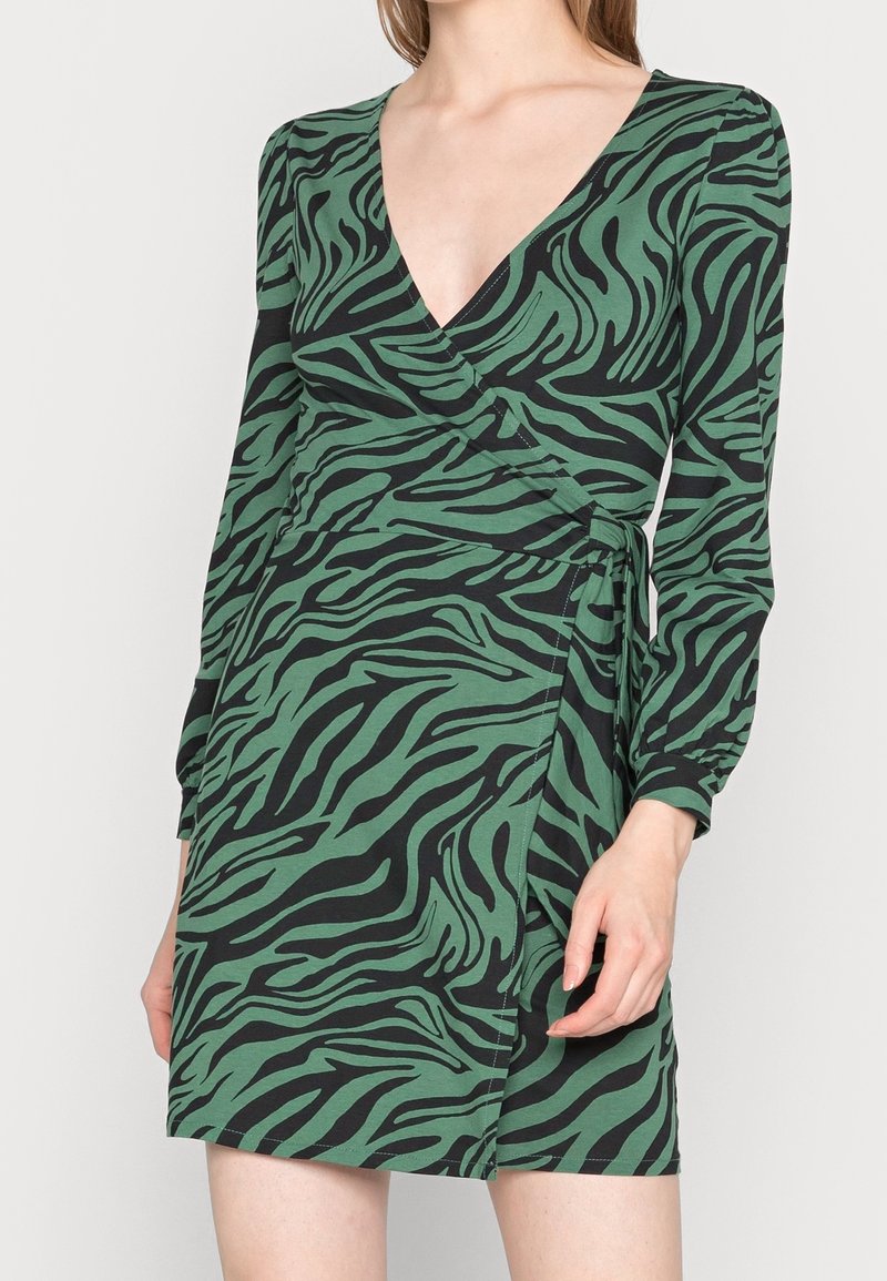 Femme portant une robe portefeuille à manches longues et à encolure en V, imprimée zèbre en vert et noir.