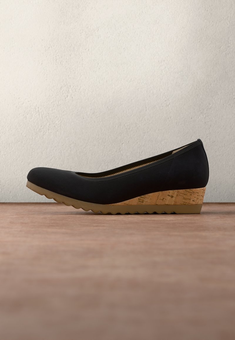 Chaussure à talon compensé noire avec talon en liège et semelle marron clair rainurée, posée sur une surface en bois devant un mur blanc texturé.