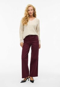 Maglione lavorato a maglia beige con scollo a V, abbinato a pantaloni larghi bordeaux. Scarpe col tacco nere completano l'outfit. Texture liscia in tutto.