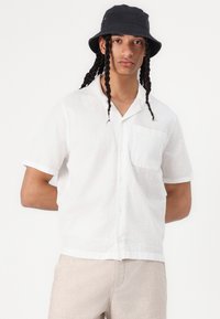 Chemise boutonnée blanche à manches courtes avec un motif texturé, dotée d'une poche poitrine et d'une coupe décontractée, associée à un pantalon beige clair.