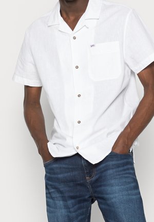 Homme portant une chemise blanche à manches courtes avec poche et un jean bleu, les mains dans les poches, sur un fond uni.