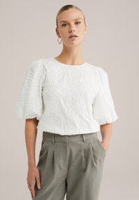 Žena v bílé texturované blůze s krátkým rukávem a nabíranými rukávy, v šedých kalhotách s vysokým pasem a záhyby, stojí s jednou rukou v kapse.