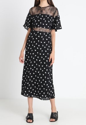 Czarna sukienka midi z białym kwiatowym wzorem, przezroczystym koronkowym karczkiem i falbaniastymi rękawami. Luźna tkanina i swobodna sylwetka. Noszona z czarnymi sandałami.