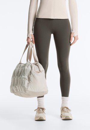 Person wearing tight dark leggings, beige long-sleeve top, white socks, beige sneakers, holding a light beige quilted duffel bag by side.