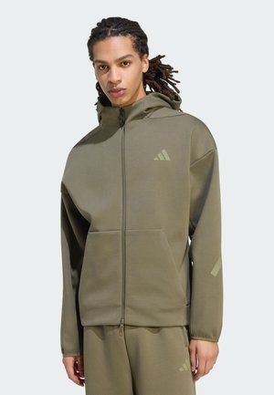 Person iført olivengrøn hættejakke med lynlås og matchende bukser med subtil adidas-logo på bryst og ben, stående mod ensfarvet baggrund.