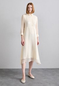 Frauen stehen in einem cremefarbenen Midikleid mit langen Ärmeln, Knöpfen und einem Kragen, tragen beigen Ballerinas und stehen vor einem schlichten grauen Hintergrund.