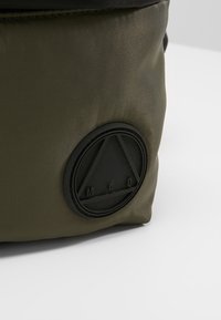 Borsa in nylon verde oliva con un patch in gomma nera testurizzata che presenta un design triangolare e le lettere "M.C.O." stampate su di essa.