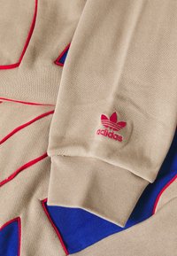Beige mikina má červené a modré akcenty, texturovanou pletenou látku a malou růžovou značku Adidas na manžetě.