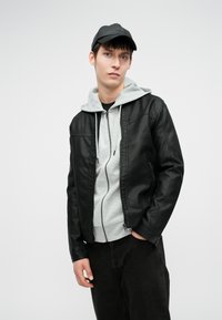 Giacca di pelle nera sopra un felpa con cappuccio grigia chiara, indossata con jeans scuri. La giacca ha una chiusura con zip e sottili dettagli di cucitura.