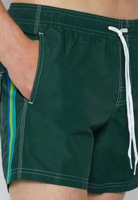 Costumi da bagno verde scuro con vita elastica e cordino bianco. Tasche laterali con strisce blu e verdi a contrasto.