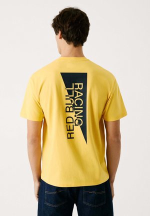 Gul bomulds T-shirt med et stort sort grafisk motiv med teksten "RED BULL RACING" i et diagonalt layout på ryggen. Korte ærmer og rund hals.