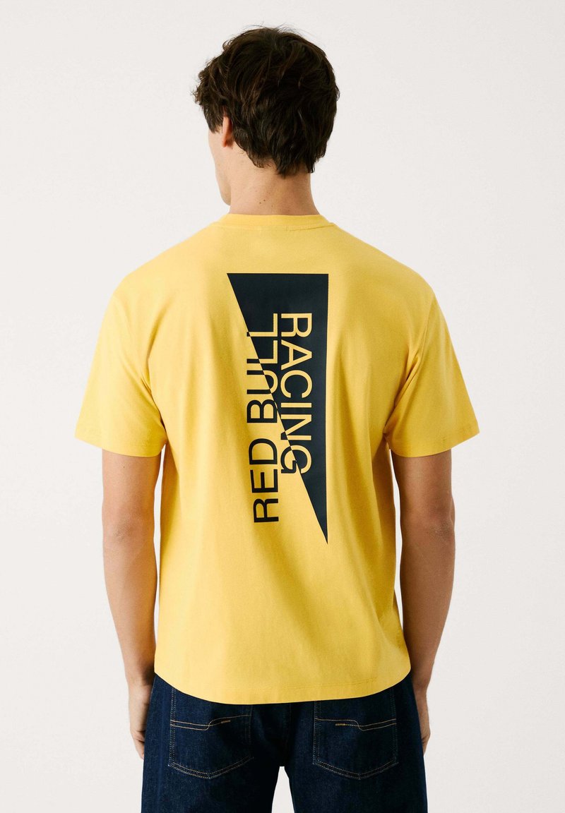 Gul bomulds T-shirt med et stort sort grafisk motiv med teksten "RED BULL RACING" i et diagonalt layout på ryggen. Korte ærmer og rund hals.
