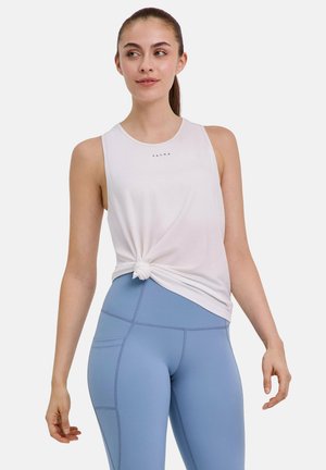 Giovane donna che indossa una canottiera bianca annodata in vita e leggings azzurri a vita alta, in piedi con le braccia rilassate.