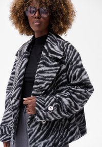 Zwart en grijs gestreepte nepbontjacket met een geknikte kraag, knopen aan de voorkant en zijzakken, gedragen over een zwarte top.